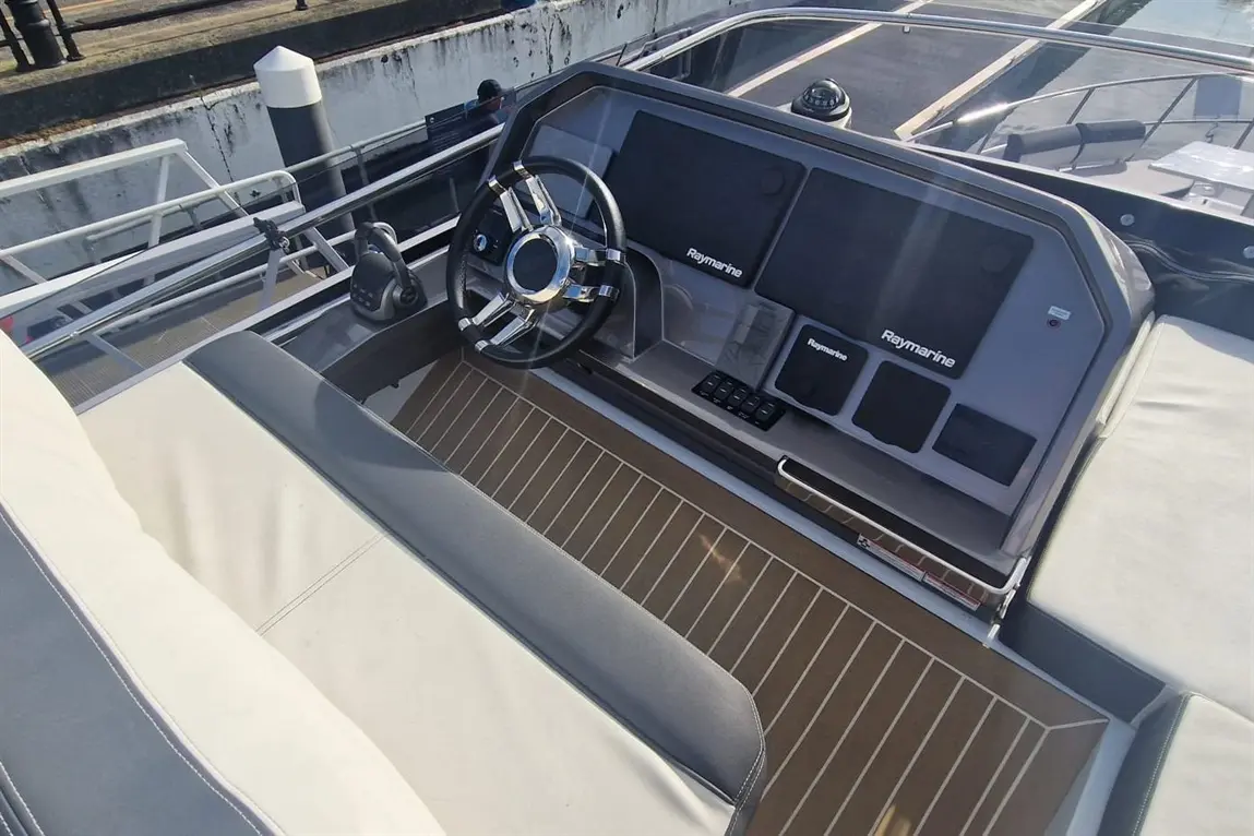 Boat helm with a Raymarine dashboard, steering wheel, control panel, and seating in a sunny marina setting.
